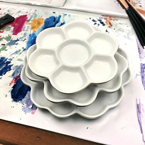 May include: Three white ceramic paint palettes stacked on top of each other. Each palette has a flower-shaped design with eight wells for mixing paint.