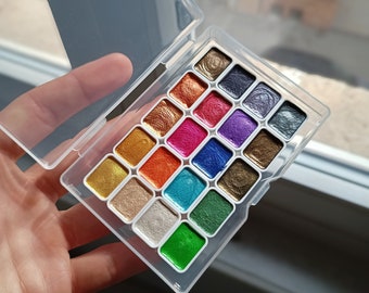 May include: A white plastic palette with 18 round paint pans. The pans are filled with metallic watercolor paints in various colors, including gold, silver, green, blue, red, and purple.
