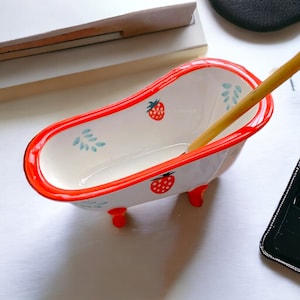 May include: A white ceramic bathtub with red trim and strawberry designs. The bathtub has four small feet and is empty.