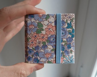 May include: A small, floral patterned notebook with a blue elastic band closure. The notebook has a white background with blue and pink flowers.