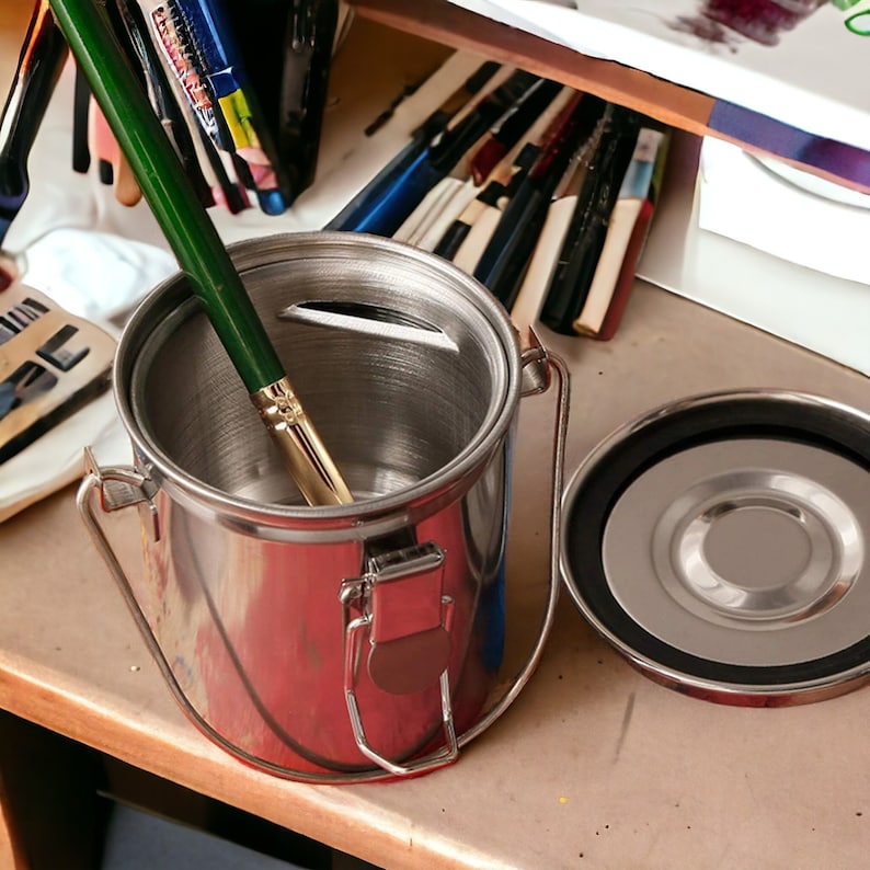 May include: A stainless steel paint brush cleaning container with a lid and a green paint brush inside. The container has a handle and a latch.