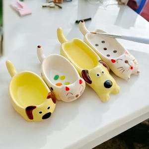 Op de afbeelding: Vier keramische verfpaletten in de vorm van katten en honden met gele en witte verf. Elke palet heeft een klein, ondiep bakje om verf in te bewaren.