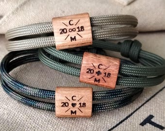 May include: Three personalized bracelets with wooden rectangular clasps. The bracelets feature braided cord in shades of green, blue, and gray. Each clasp is engraved with initials, a date, and a heart symbol, creating a custom accessory.