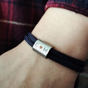 May include: A navy blue bracelet with a silver clasp. The clasp is engraved with the letters "C", a heart symbol, and "M". The bracelet is made of multiple strands of navy blue cord.