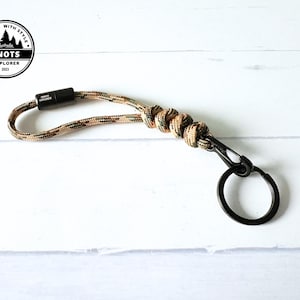 May include: Camouflage paracord keyring with a black metal clasp and ring. The keyring has a braided design in shades of brown, green, and tan. The Knots Explorer logo is in the upper left corner.