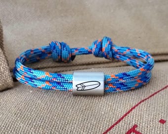 May include: A blue, orange, and purple patterned cord bracelet featuring a silver-toned metal bead. The bead is engraved with a black line drawing of a surfboard. The bracelet is constructed with two adjustable sliding knots for a secure fit.