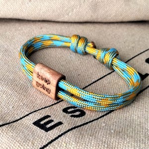 May include: A blue and yellow braided bracelet with a wooden clasp. The clasp is engraved with the words "keep going". The bracelet is on a tan fabric surface.