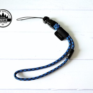 Op de afbeelding: Een blauw en zwart patroon polsbandje met een zwarte gesp en een klein zwart leren label met de tekst "CHAMP". De band heeft een lus aan het uiteinde en een zwarte plastic sluiting. Het product ligt op een wit houten oppervlak.