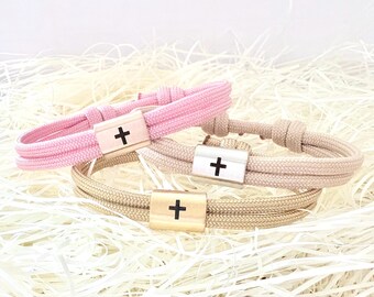 May include: Three rope bracelets in pink, tan, and gold. Each bracelet features a wooden or silver-toned clasp with a black cross design. The bracelets are displayed on a bed of white shredded paper.