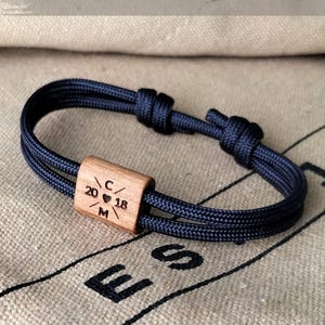May include: A navy blue rope bracelet with a wooden centerpiece. The wooden piece is engraved with the numbers "20 18", a heart symbol, and the letters "C" and "M". The bracelet is knotted for adjustment.