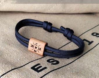 May include: A navy blue rope bracelet with a wooden centerpiece. The wooden piece is engraved with the numbers "20 18", a heart symbol, and the letters "C" and "M". The bracelet is knotted for adjustment.