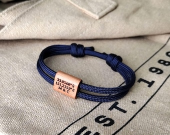 May include: A navy blue bracelet with a wooden rectangular bead. The bead is engraved with coordinates and the initials "M & C". The bracelet is made of two parallel cords, knotted at each end.