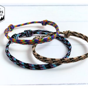 May include: Three adjustable cord bracelets featuring woven patterns. One bracelet has a vibrant multi-coloured design, another is black with purple and blue accents, and the third is a tan and brown camouflage pattern. A logo reads KNOTS EXPLORER Australia.