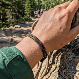 May include: A multi-strand bracelet made of thin, braided cord featuring a dark, mottled pattern of black, green, and brown tones. The bracelet is worn on the wrist, showcasing a textured, woven design that wraps around multiple times.