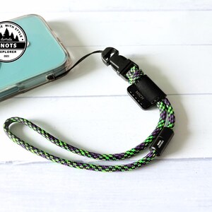 Op de afbeelding: Een telefoon polsbandje met een zwarte sluiting en een groen, paars en zwart touwontwerp. De band is bevestigd aan een doorzichtige telefoonhoes. De afbeelding bevat een logo met de tekst "Explore with Style" en "Knots Explorer 2023".
