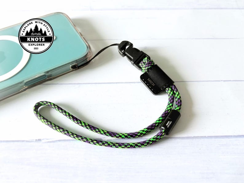 Op de afbeelding: Een telefoon polsbandje met een zwarte sluiting en een groen, paars en zwart touwontwerp. De band is bevestigd aan een doorzichtige telefoonhoes. De afbeelding bevat een logo met de tekst "Explore with Style" en "Knots Explorer 2023".