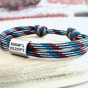 May include: A multi-strand paracord bracelet featuring a cylindrical silver-toned metal bead engraved with the coordinates 33.8568 S, 151.2153 E. The cord is woven with a repeating pattern of black, white, red, and teal blue threads and includes two sliding knots.