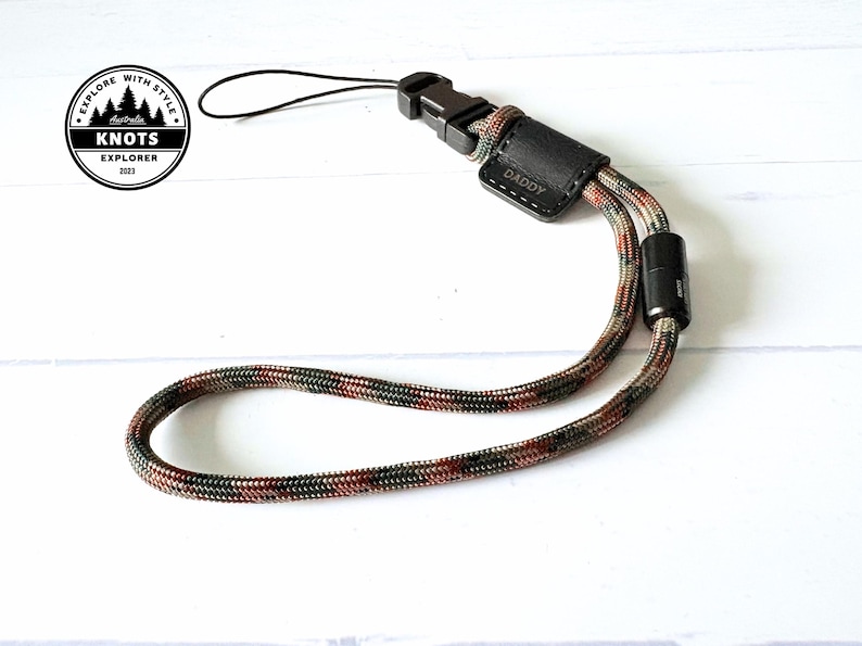 Op de afbeelding: Een polsband met een camouflagepatroon in bruin, groen en rood. Het heeft een zwarte plastic gesp en een klein leren stukje met het woord "DADDY" in reli&euml;f. De band is ontworpen voor het vastzetten van items. Lengte ongeveer 20 cm.