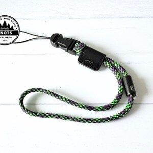 Op de afbeelding: Een zwart-groene polsband met een zwarte gesp en een klein zwart leren label met de tekst "MICH". De band is gemaakt van een geweven koord met groene, paarse en zwarte kleuren. Het product ligt op een wit oppervlak.