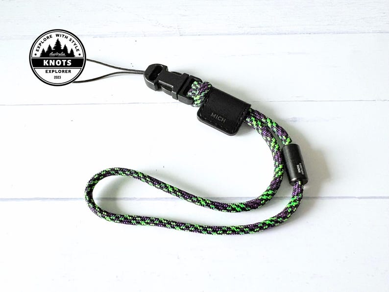 Op de afbeelding: Een zwart-groene polsband met een zwarte gesp en een klein zwart leren label met de tekst "MICH". De band is gemaakt van een geweven koord met groene, paarse en zwarte kleuren. Het product ligt op een wit oppervlak.