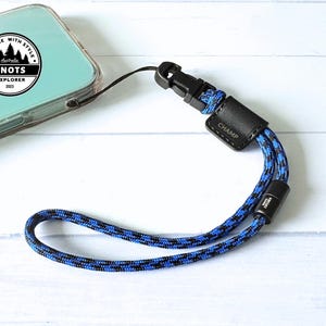 Op de afbeelding: Een blauw en zwart touw polsbandje voor de telefoon met een zwarte plastic sluiting en een zwart leren label met de tekst "CHAMP". De telefoon zit in een doorzichtige hoes. De band is aan de telefoon bevestigd. De afbeelding bevat ook een logo met de tekst "EXPLORE WITH STYLE" en "KNOTS EXPLORER 2023".