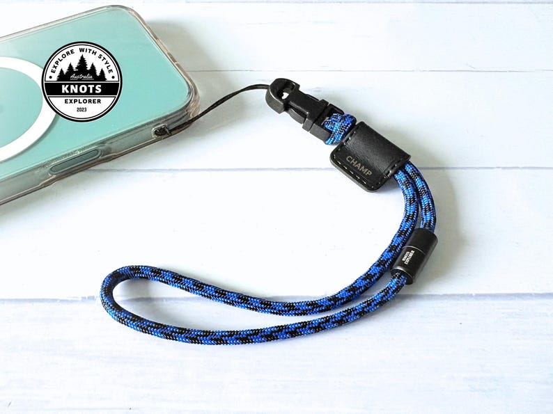 Op de afbeelding: Een blauw en zwart touw polsbandje voor de telefoon met een zwarte plastic sluiting en een zwart leren label met de tekst "CHAMP". De telefoon zit in een doorzichtige hoes. De band is aan de telefoon bevestigd. De afbeelding bevat ook een logo met de tekst "EXPLORE WITH STYLE" en "KNOTS EXPLORER 2023".