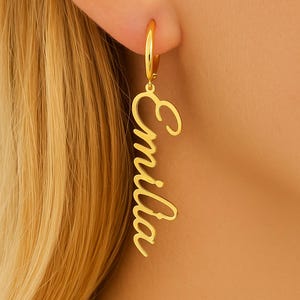 Può includere: Orecchino a cerchio color oro con il nome "Emilia" in corsivo. L'orecchino è appeso verticalmente, con il nome scritto dall'alto verso il basso. L'orecchino è attaccato a un cerchio d'oro che è attaccato all'orecchio.