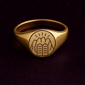 May include: A gold-colored signet ring with a circular emblem featuring a mountain range, sun rays, and two tablets. The ring has a smooth, polished finish and a classic, rounded shape. The ring is a size 9.