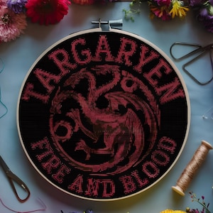 Puede incluir: Un bastidor de bordado de punto de cruz con el emblema Targaryen y el texto "FIRE AND BLOOD" en rojo sobre fondo negro. El bordado está en un bastidor de madera, rodeado de flores coloridas y materiales de manualidades.