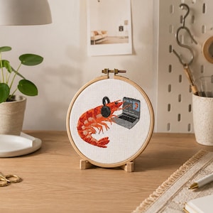May include: A circular wooden embroidery hoop containing white fabric with a cross-stitch design of a bright orange shrimp wearing black headphones while sitting in front of a small grey laptop computer. The shrimp is facing the laptop screen.