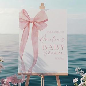 May include: A baby shower welcome sign on a wooden easel. The sign features a large, watercolor-style pink bow and the words "Welcome to Amelia's BABY shower" in elegant script. The date is also included.