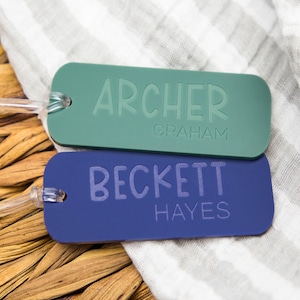 May include: Two rectangular luggage tags, one teal and one blue, with clear plastic loops. The teal tag reads "ARCHER" over "GRAHAM." The blue tag reads "BECKETT" over "HAYES." The tags are on a striped cloth and a woven surface.