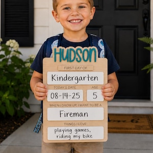 May include: A light-colored wooden sign shaped like a clipboard with the name Hudson in teal script at the top. The sign features text fields for First Day of Kindergarten, date 08-14-25, age 5, career goal Fireman, and interests playing games, riding my bike.