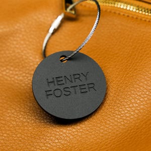 May include: A black round tag with the text "HENRY FOSTER" attached to a brown leather bag with a gold zipper.