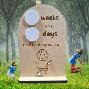 May include: A wooden countdown board with two blank circles for weeks and days. The board says "weeks and days until I get my cast off". A cartoon drawing of a person with a cast on their leg is below the circles. The board is in front of a green grassy area with trees in the background.