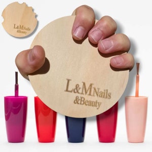 Puede incluir: Cinco botellas de esmalte de uñas en diferentes colores, incluyendo rosa, rojo, azul y morado. Las botellas están dispuestas en una fila sobre una superficie blanca. Un círculo de madera con el texto "L&M Nails & Beauty" está siendo sostenido por una mano.
