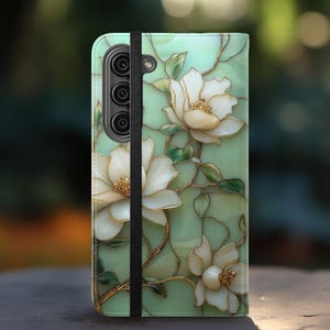 May include: A mint green phone case featuring a floral design with white flowers and gold accents. The case has a black elastic band closure and a camera cutout. The floral pattern is reminiscent of stained glass.