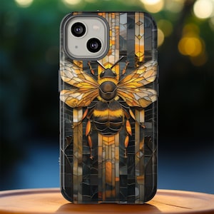 Può includere: Una custodia per telefono nera con un design a vetri colorati che presenta un'ape con strisce gialle e nere. L'ape è circondata da un mosaico di pezzi dorati, neri e bianchi.