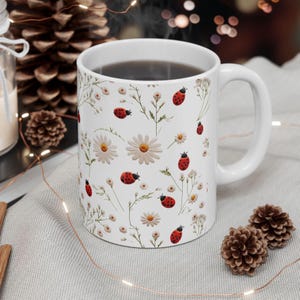 May include: White ceramic mug with a floral pattern featuring white daisies and red ladybugs. The mug is filled with dark coffee.