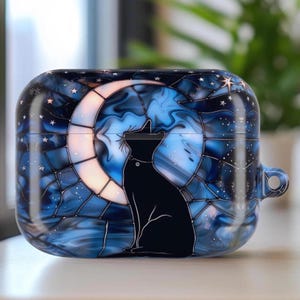 May include: AirPods case with a celestial design. The case features a black cat silhouette sitting in front of a crescent moon. The background is a swirling blue and white pattern with stars. The case has a glossy finish and a small loop for attaching a lanyard.