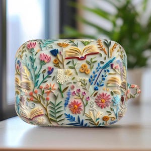 May include: A white AirPod case decorated with a vibrant floral and book pattern. The case showcases various flowers in shades of pink, orange, blue, and yellow, alongside open book illustrations. The design is intricate and textured, offering a distinctive look.