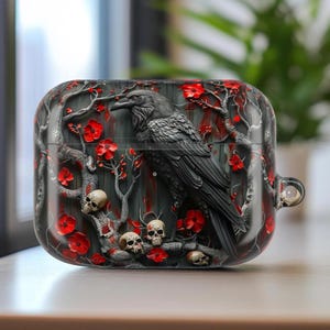 May include: A black and gray AirPod case featuring a detailed raven design. The case has a dark gray background with red flowers, tree branches, and small skulls. The raven is perched on a branch, adding a gothic aesthetic.