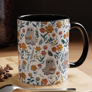 May include: White ceramic mug with a black interior and handle. The mug features a repeating pattern of smiling cartoon ghosts holding flowers, set against a backdrop of orange, yellow, and pink flowers and green foliage.