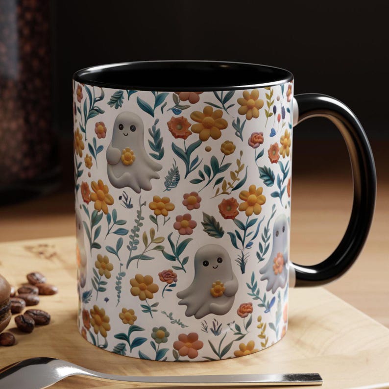 May include: White ceramic mug with a black interior and handle. The mug features a repeating pattern of smiling cartoon ghosts holding flowers, set against a backdrop of orange, yellow, and pink flowers and green foliage.