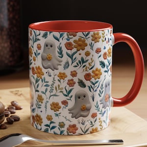 May include: A ceramic mug with a red interior and handle. The exterior features a white background with a floral pattern and cartoon ghosts holding flowers. The mug is sitting on a wooden surface.