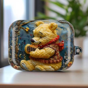 May include: A yellow Winnie the Pooh-themed case for wireless earbuds. The case features a 3D design of the bear holding a basket of honey, wearing a red scarf and a brown sweater. The background is a blue and green landscape.