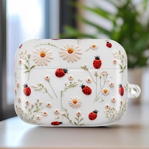 May include: White AirPod case decorated with embossed daisies, small white flowers, green stems, and red ladybugs. The case has a smooth, glossy finish and a small loop for attaching a lanyard.