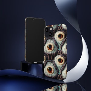 May include: Two phone cases, one black and one with a pattern of embroidered eyes in shades of blue, brown, and white.