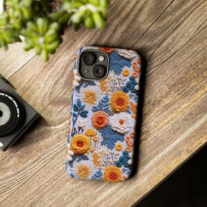 Pode incluir: Uma capa de telefone com um design floral com flores amarelas, laranja, brancas e azuis sobre um fundo azul claro. A capa tem um recorte para a c&acirc;mera com tr&ecirc;s lentes.