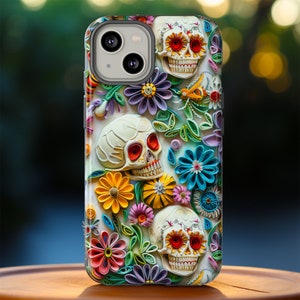 May include: A phone case with a colorful floral design featuring three white sugar skulls with black eyes and red accents. The case has a white background with a variety of colorful flowers and leaves.